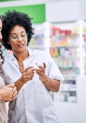 Woman in a pharmacy talking to a female pharmacist and looking at a bottle of medication the pharmacist is holding. 