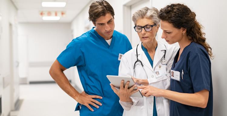 Mature female doctor discussing medical report with nurses in hospital hallway. Senior general practitioner discussing patient case status with group of medical staff after surgery. Doctor working on digital tablet while in conversation with healthcare workers, copy space.