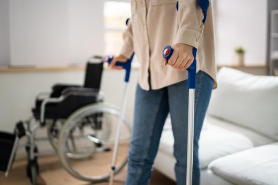 A mid shot of an individual using crutches in their home, in the background there is a wheelchair