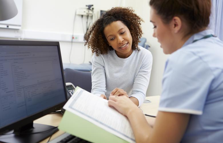 Two women are in a clinical room. One is looking in the direction of the camera. She is the patient. The other is in side profile. She is the doctor. They are looking at a chart together. A computer monitor is on the left of the image.