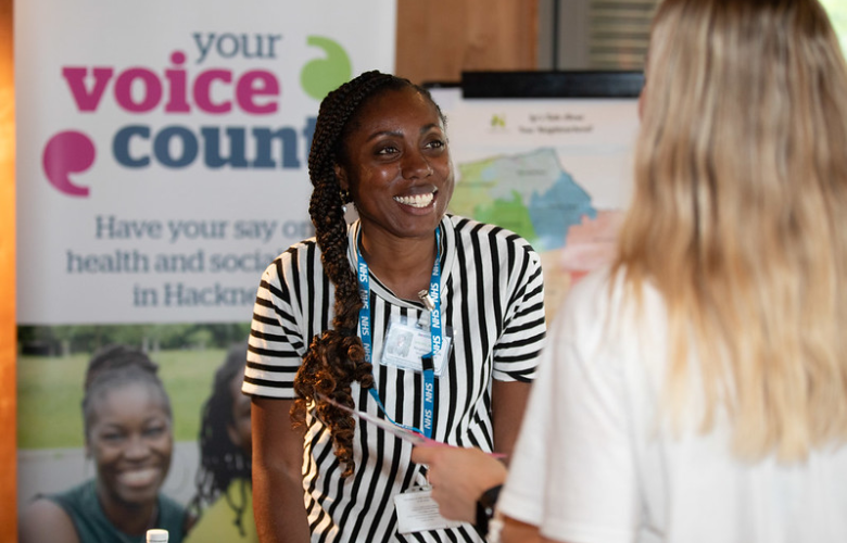 A woman is standing at an information booth and smiling. She is speaking to another woman, whose back is to the camera. In the background a Healthwatch branded sign saying 'your voice counts'